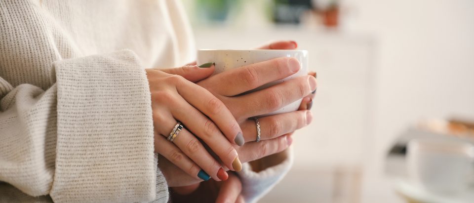Two pairs of hands in knitted sleeves closed around a mug held between them.
