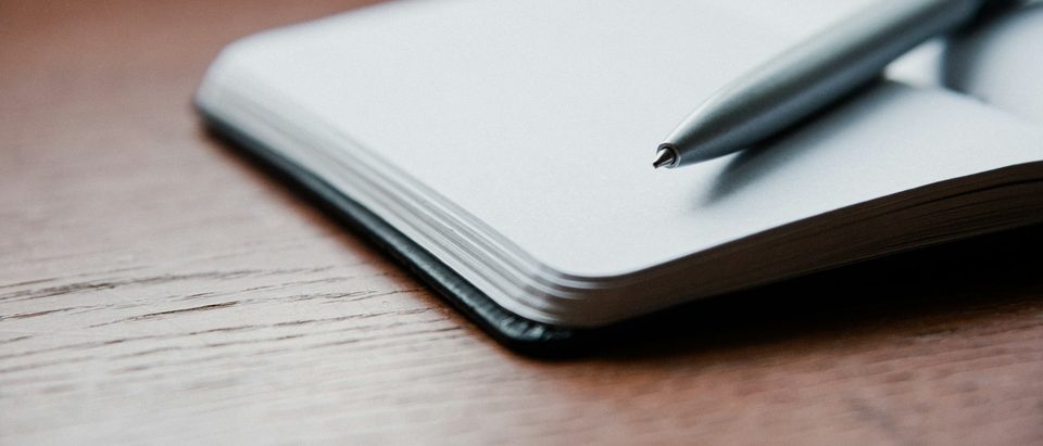 An open notebook and a pen lying on a wooden table in low morning light.
