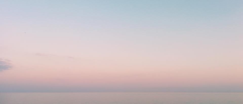 Still sea water under a flat dawn sky, pink and grey where the light meets the horizon.